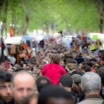 A crowd of people at El Rastro market on Calle Embajadores in Madrid. On either side are market stalls with goods for sale, with trees lining the street in the background and a child sitting on someone's shoulders with his back to the camera in the foreground.
