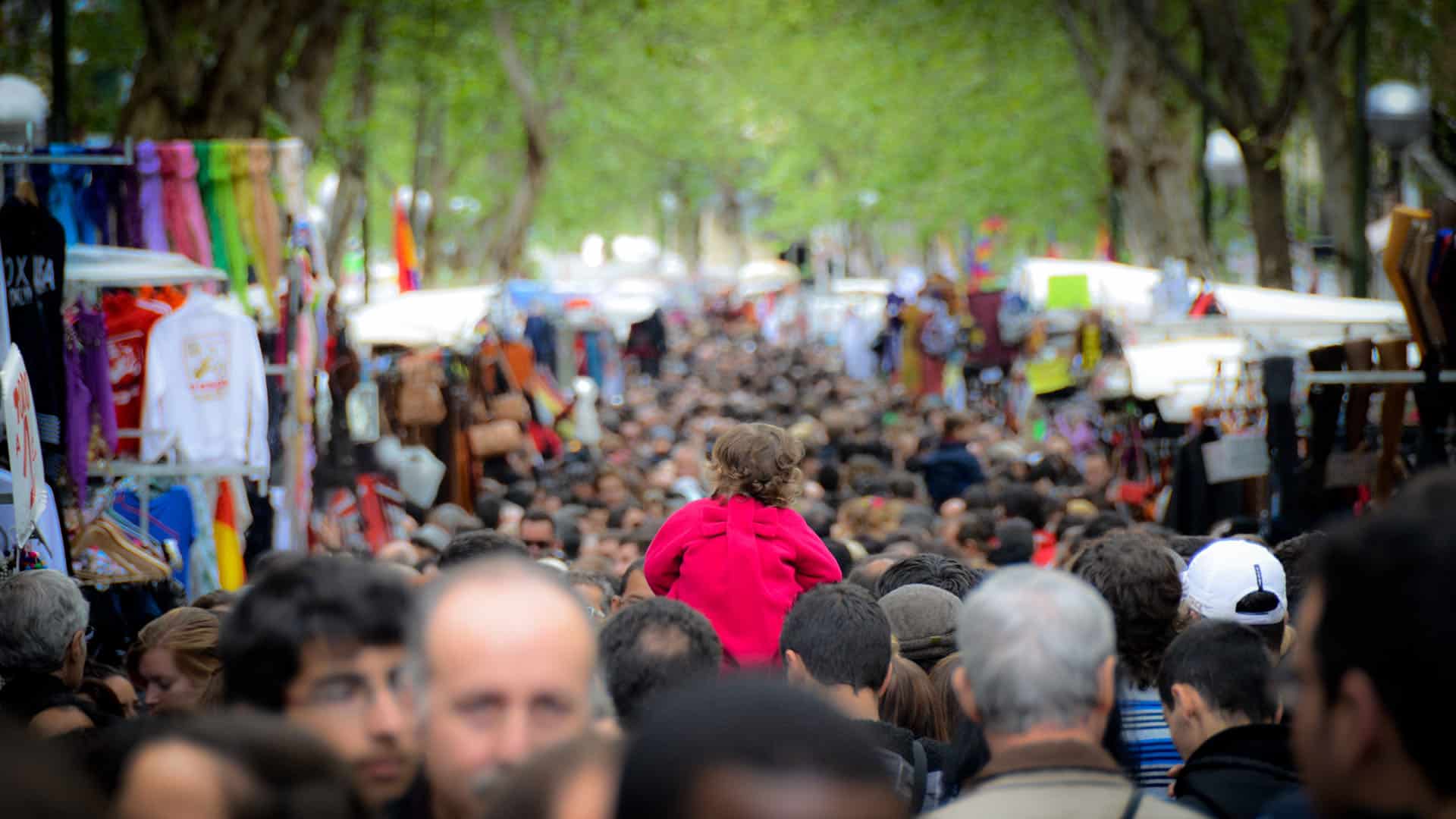 A crowd of people at El Rastro market on Calle Embajadores in Madrid. On either side are market stalls with goods for sale, with trees lining the street in the background and a child sitting on someone's shoulders with his back to the camera in the foreground.