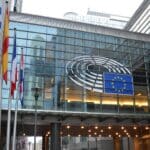 Imagen de una de las entradas al edificio del Parlamento Europeo en Bruselas. La fachada de vidrio despliega el logo del Parlamento y la bandera de la Unión Europea. A la izquierda del edificio gris, algunas banderas de los estados miembros de la UE.
