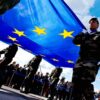 Grupo de militares sosteniendo una gran bandera azul con estrellas amarillas, símbolo de la Unión Europea, durante una ceremonia al aire libre frente al Parlamento Europeo, en la primera sesión plenaria del Parlamento Europeo que marca el inicio para el mandato 2014-2019. Al fondo el cielo parcialmente nublado y una multitud tras una valla observando.