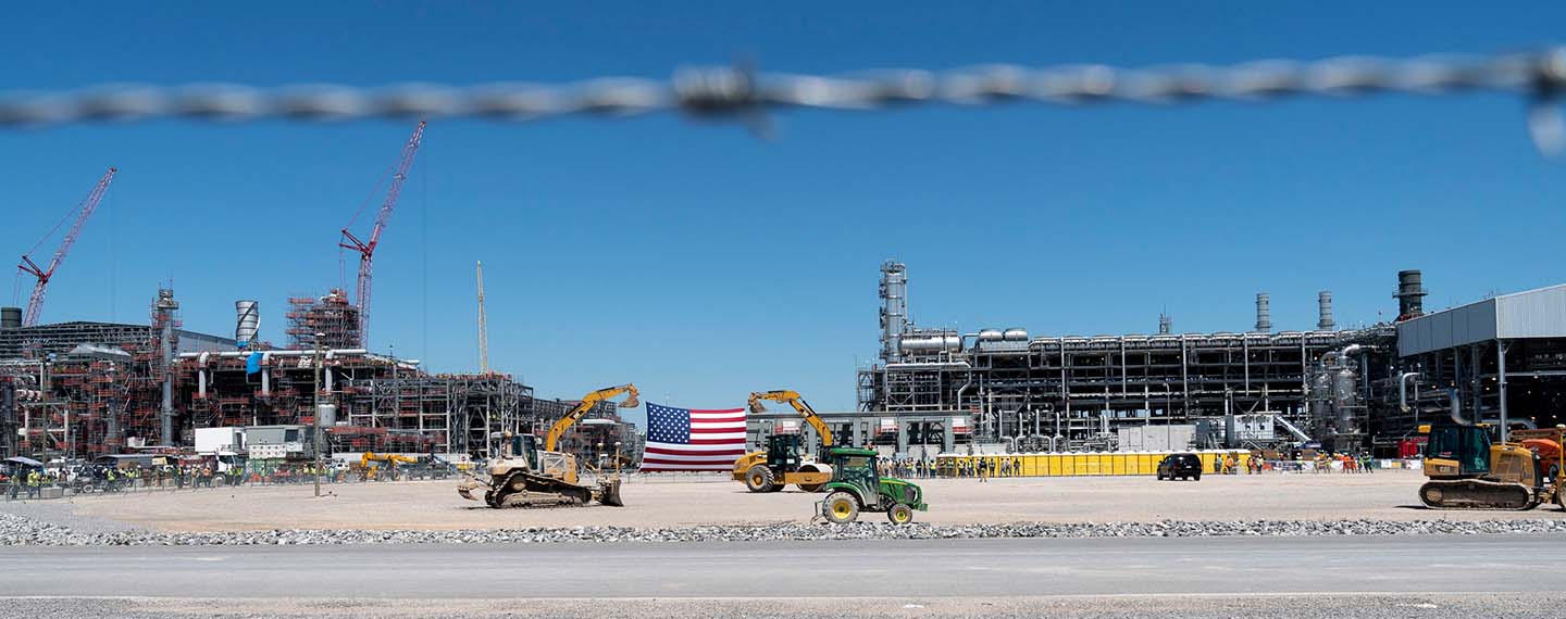 Foto tomada a través de una alambrada de la terminal de exportación en la planta de licuefacción de gas natural licuado Cameron en Hackberry, Louisiana (EEUU). En el centro se observa una bandera estadounidense gigantesca en medio de dos maquinarias de palas de construcción