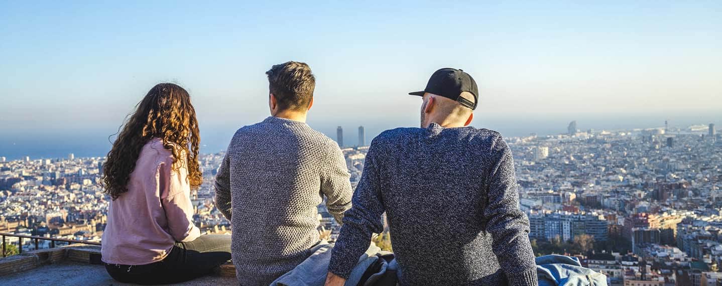 Three young adults friends sitting on a wall overlooking the city of Barcelona, Spain Three young adults (one woman and two men) sitting on a stone wall looking at the skyline of the city of Barcelona