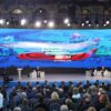 Reunión del presidente ruso Vladimir Putin con más de 500 partidarios oficiales en el Gostiny Dvor en Moscú antes de las elecciones presidenciales de 2024. La sala reúne una multitud de partidarios, trabajadores del evento y medios de comunicación. En el escenario, dos pantallas a cada lado muestran la imagen de Putin y una pantalla central proyecta la imagen del evento. Putin está sentado a la izquierda y a la derecha los tres moderadores del evento