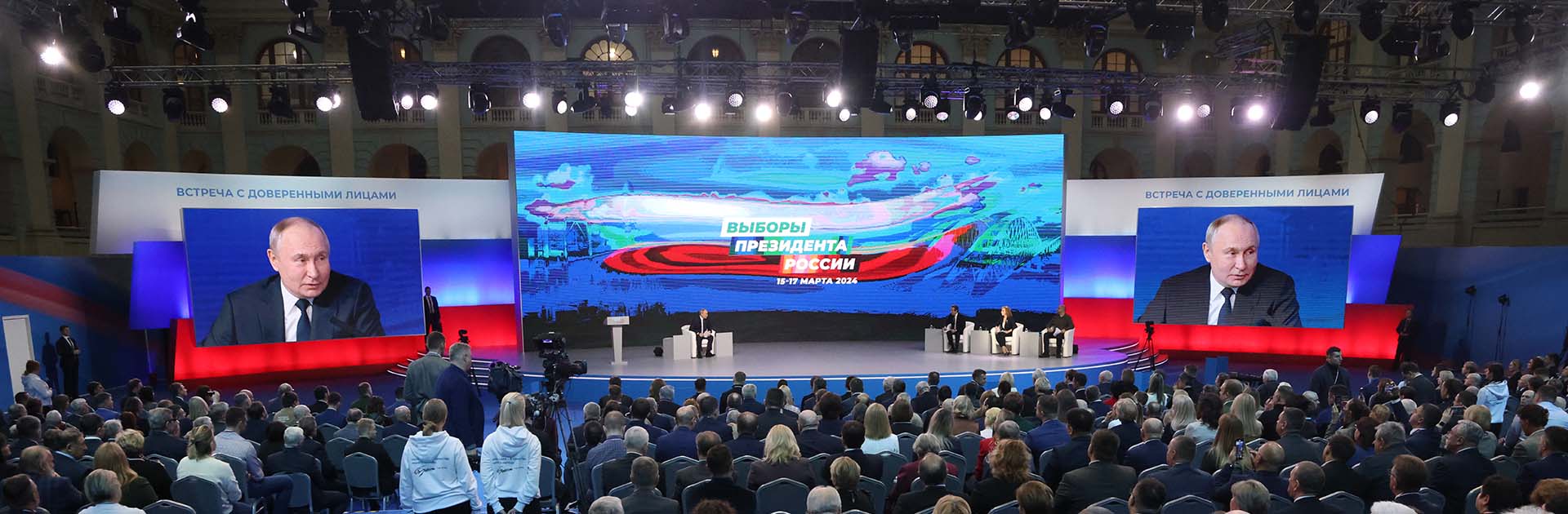 Reunión del presidente ruso Vladimir Putin con más de 500 partidarios oficiales en el Gostiny Dvor en Moscú antes de las elecciones presidenciales de 2024. La sala reúne una multitud de partidarios, trabajadores del evento y medios de comunicación. En el escenario, dos pantallas a cada lado muestran la imagen de Putin y una pantalla central proyecta la imagen del evento. Putin está sentado a la izquierda y a la derecha los tres moderadores del evento