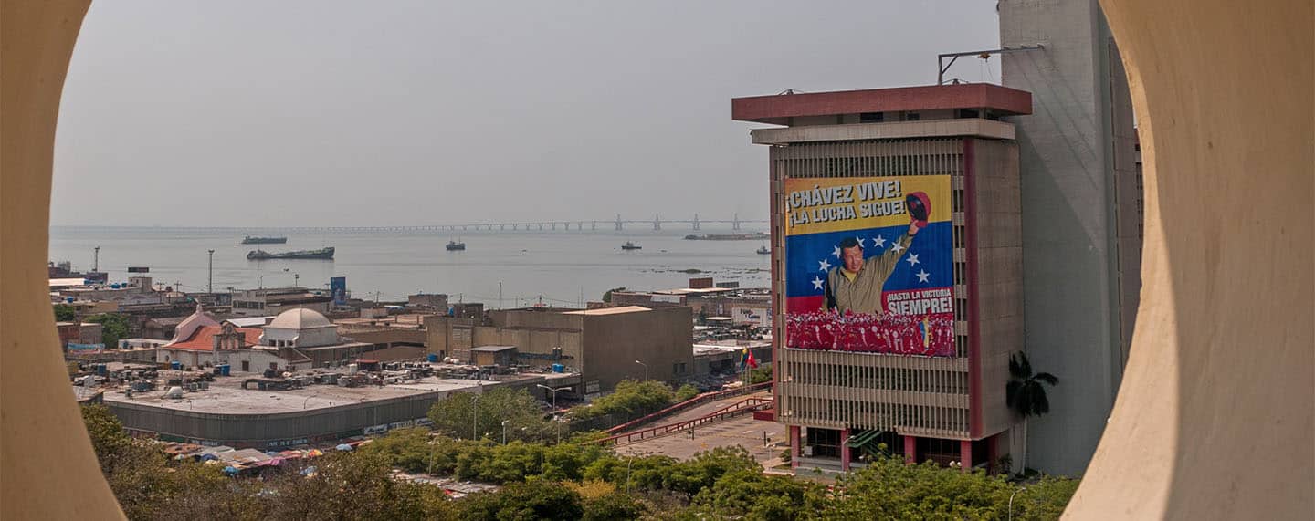 Vista de las Torres de PDVSA desde el campanario de la Basílica de la Virgen de Chiquinquirá, en la ciudad de Maracaibo (Venezuela). En la torre en primer plano cuelga un cartel de propaganda del gobierno, con el lema "¡Chávez vive! ¡La lucha sigue!". Al fondo, el lago de Maracaibo con buques cargueros y el puente sobre el lago