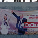 Claudia Sheinbaum y Andrés Manuel López Obrador en un cartel de propaganda electoral de MORENA en Dolores Hidalgo, Guanajuato (México). El cartel está colgado en una pared de bloques de cemento pintada de gris y amarillo. En la parte superior de la pared sobresalen trozos de madera con una alambrada