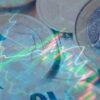 Fondo difuminado de euros en papel (20 y 10 euros) y en monedas (1 euro) y gráficos de líneas y barras que simbolizan ciclos económicos. Deuda pública