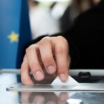 Persona depositando su voto en una urna. Fondo: Bandera de la Unión Europea.