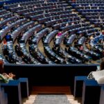Close-up of the seats in the European Parliament hemicycle, featuring microphones and electronic panels, with someone delivering her speech in the background. Europe