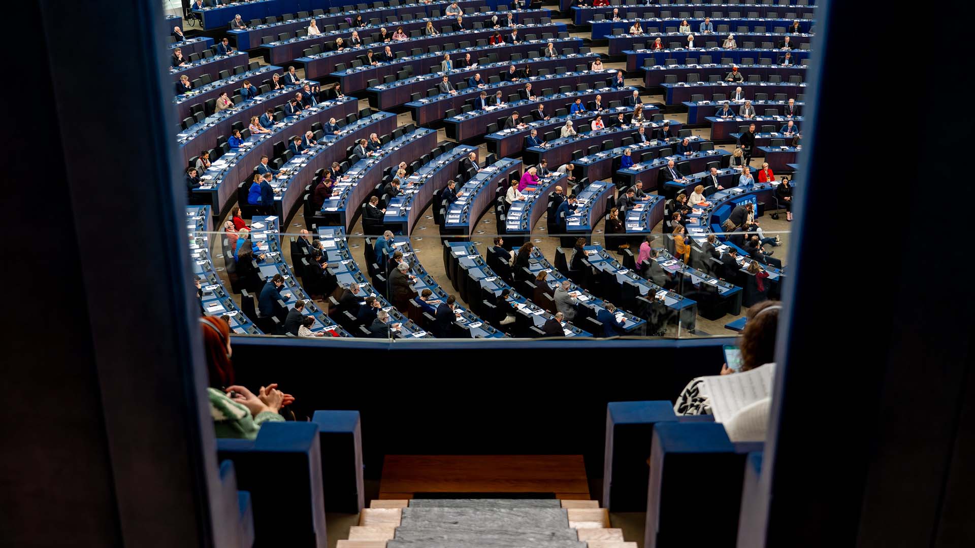 Close-up of the seats in the European Parliament hemicycle, featuring microphones and electronic panels, with someone delivering her speech in the background. Europe