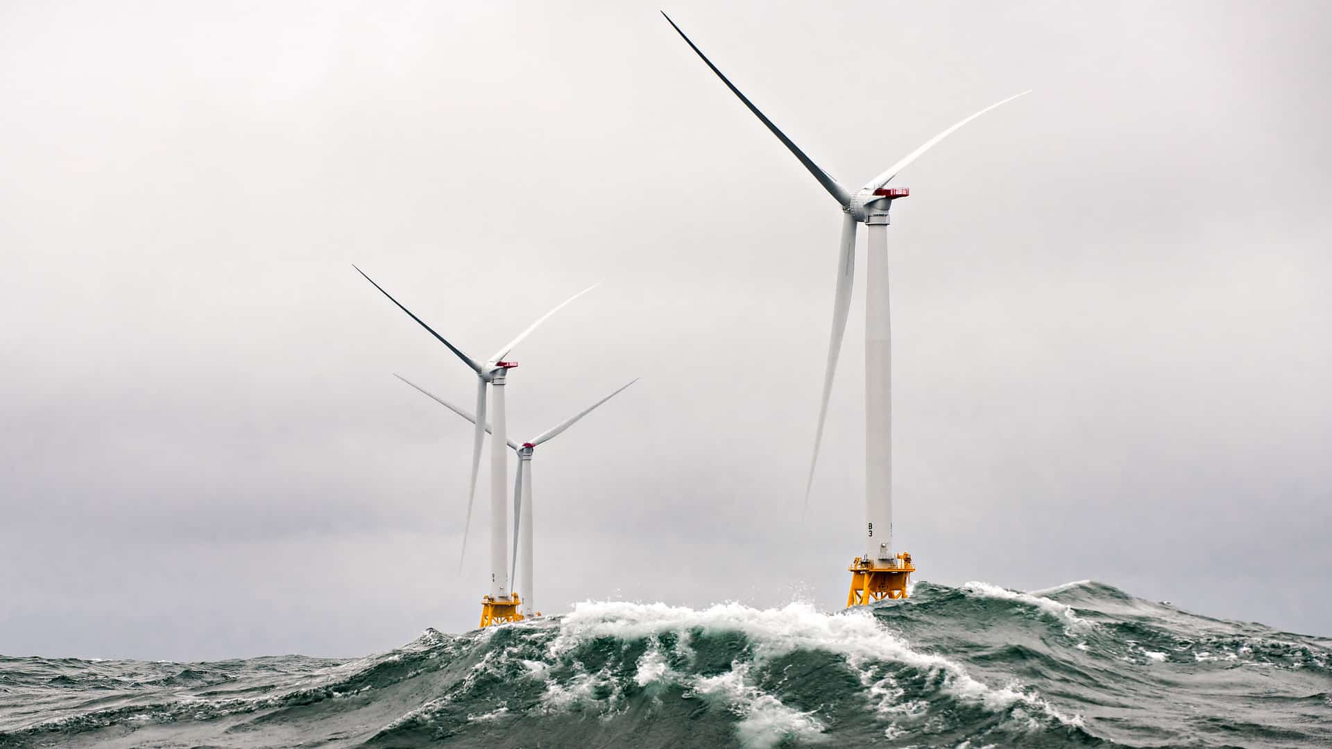 Offshore white wind turbines in rough ocean conditions, with large waves crashing against their bases under a cloudy sky. renewable energy