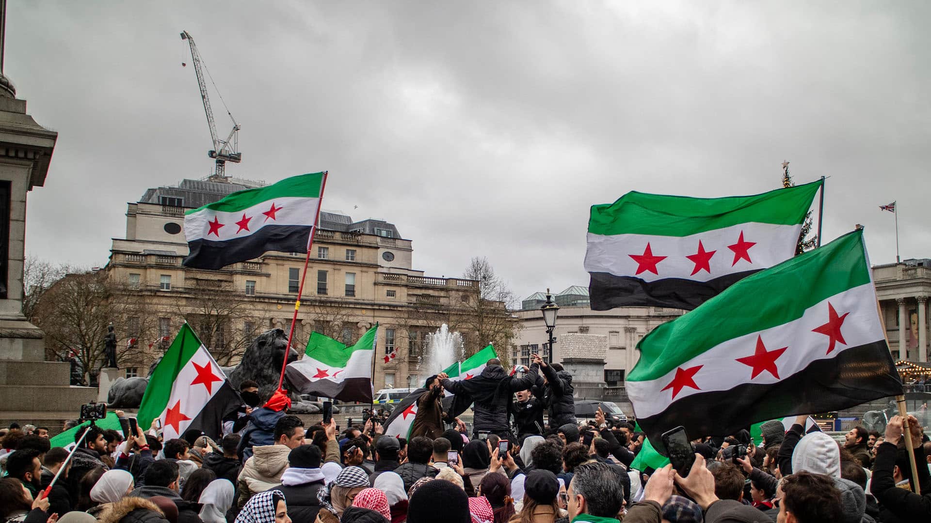 Multitud reunida en Trafalgar Square (Londres), con personas sosteniendo banderas con franjas verde, blanca y negra con tres estrellas rojas. En el fondo, se ven edificios históricos y una fuente bajo el cielo nublado. Bashar al-Assad