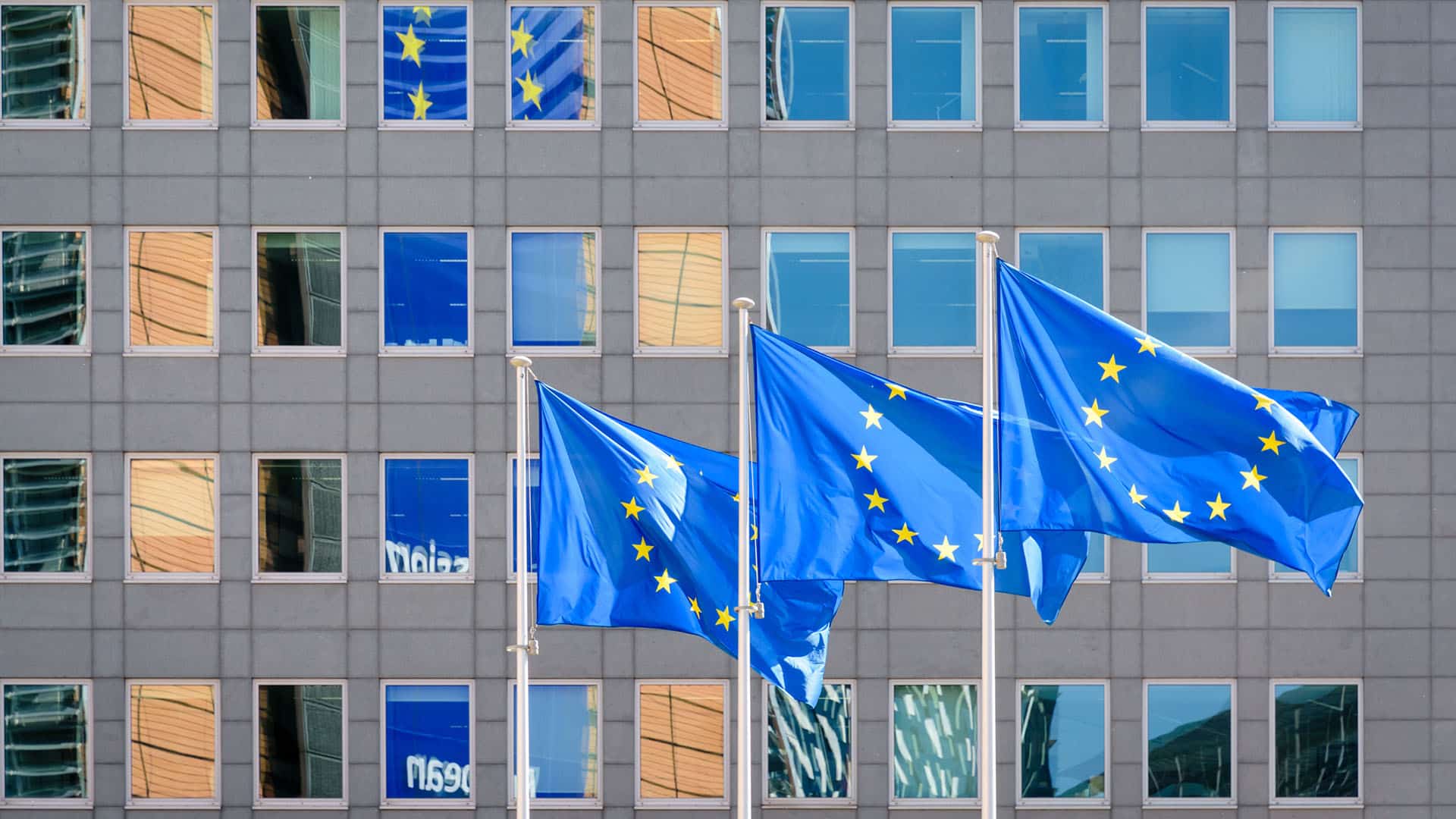 Three European Union (EU) flags fly in the wind at full mast outside the windows of the Berlaymont building, the headquarters of the European Commission (the EU executive) in Brussels, Belgium