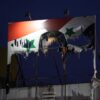 Cartel dañado de Bashar al-Assad con la bandera de Siria de fondo mostrando signos de deterioro y quemaduras, con un cielo nocturno al fondo