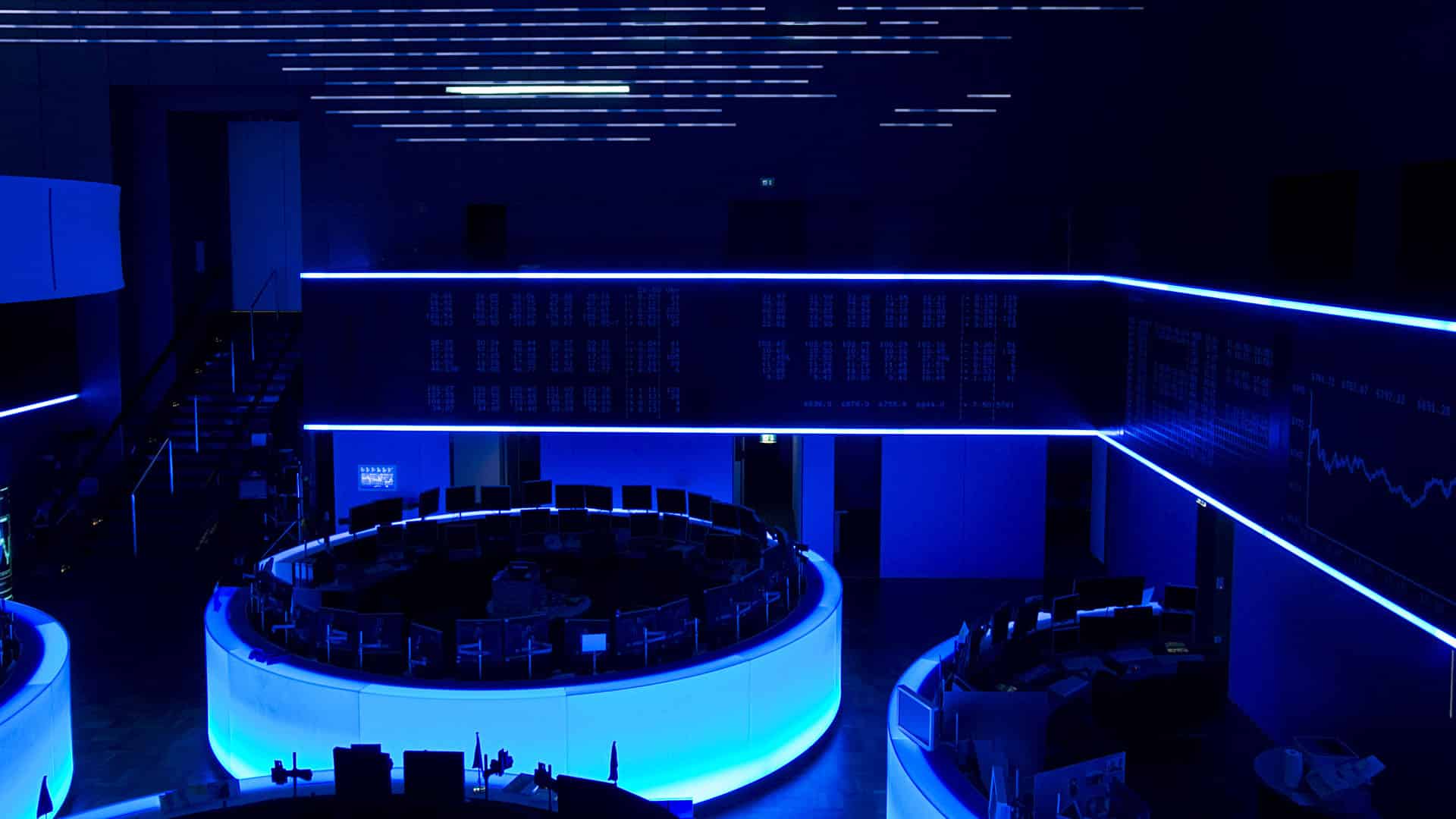 Sala de negociación en la Bolsa de Fráncfort iluminada con tonos azules, mostrando pantallas digitales y líneas de datos de mercado. Mercados financieros