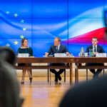 Press conference following the meeting of the Conference of Presidents of the European Parliament (EP) with the upcoming Polish Presidency of the Council of the EU. In the foreground, blurred, are television cameras and a journalist. Seated behind wooden tables (with a projection on a screen displaying the EU and Polish flags) are Roberta Metsola, President of the EP (on the left), and Donald Tusk, Prime Minister of Poland (on the right), along with members of the Polish government. EU Council