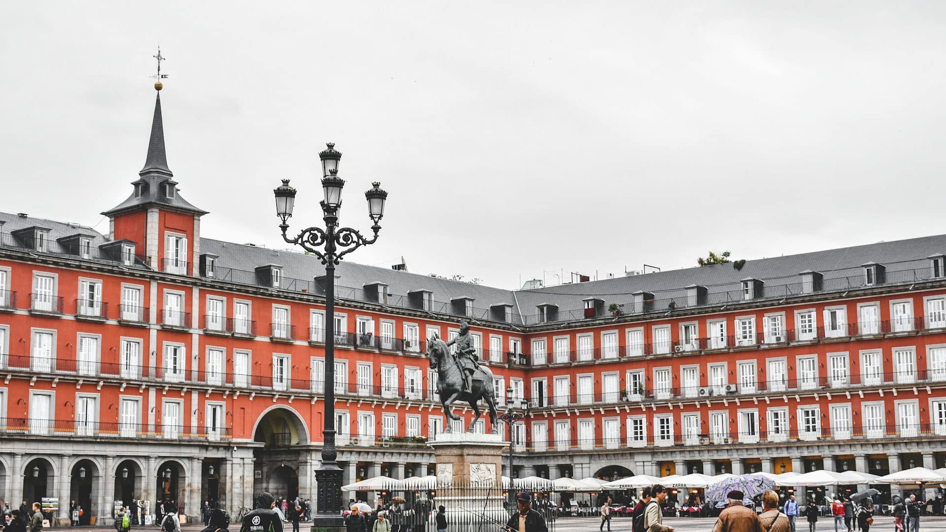 Vista de la Plaza Mayor de Madrid (España) en un día nublado. En el centro resalta la estatua ecuestre del Rey Felipe III, rodeada de edificios de fachada roja, tejados de pizarra gris y balcones alineados. Los soportales de la planta baja albergan cafés y tiendas, mientras se aprecia a varias personas caminando. En primer plano, destaca una farola negra ornamentada. Madrid