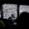 Vista aérea de una zona del este de Ucrania a través de la ventana de una aeronave en un día nublado. En primer plano, se distinguen las siluetas oscuras de dos soldados ucranianos que observan el terreno desde el interior de la cabina. En el exterior, el paisaje muestra casas dispersas con techos de chapa, calles angostas y terrenos agrícolas. Ucrania