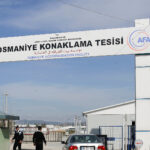 Control de seguridad en el centro de alojamiento de Osmaniye, en Turquía. En la parte superior, un cartel muestra el nombre de la instalación en tres idiomas (inglés, turco y árabe), junto con la bandera turca y el logotipo de la AFAD (Autoridad de Gestión de Desastres y Emergencias de Turquía). Debajo del cartel, un coche plateado está detenido en la entrada, mientras dos agentes de seguridad, vestidos con uniforme negro, vigilan la zona. A ambos lados, hay vallas de color azul. A la derecha, se distingue a un hombre de pie cerca de un edificio blanco. Refugiados