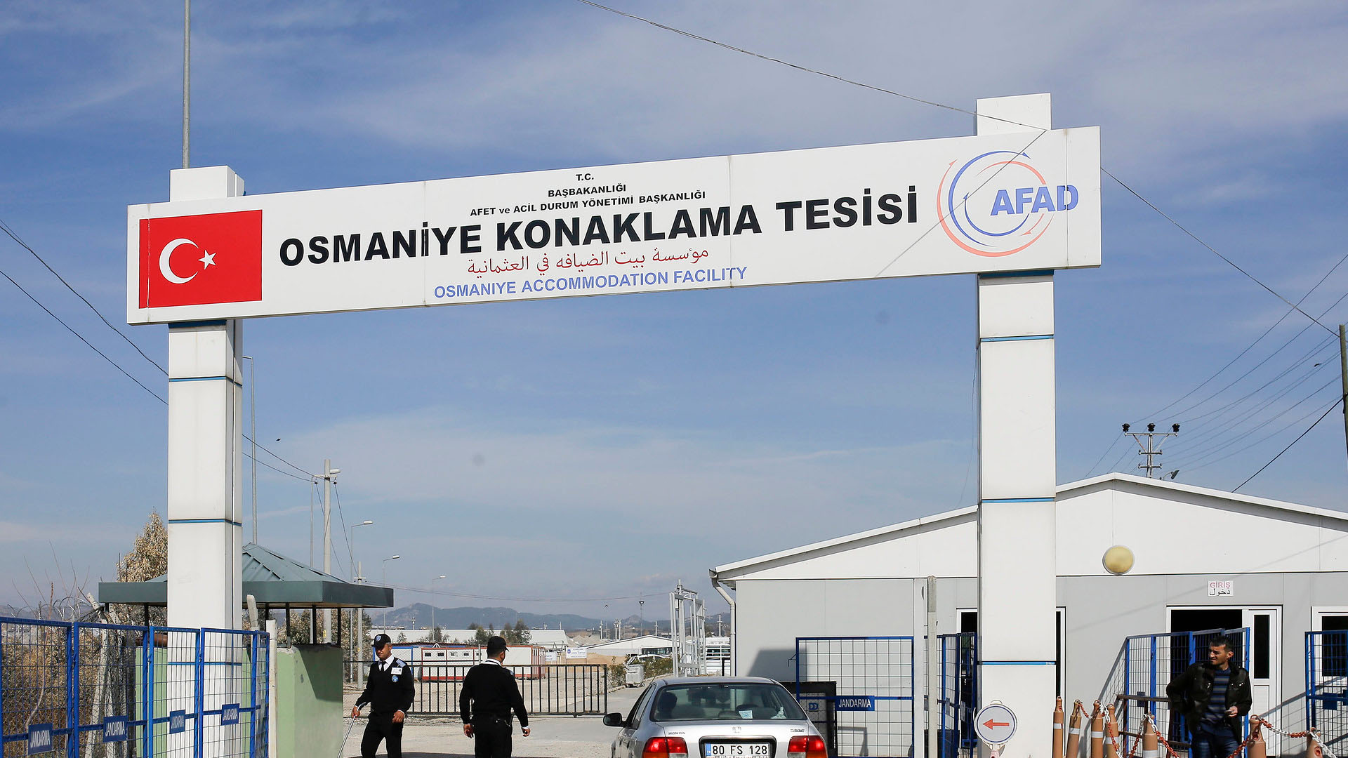 Control de seguridad en el centro de alojamiento de Osmaniye, en Turquía. En la parte superior, un cartel muestra el nombre de la instalación en tres idiomas (inglés, turco y árabe), junto con la bandera turca y el logotipo de la AFAD (Autoridad de Gestión de Desastres y Emergencias de Turquía). Debajo del cartel, un coche plateado está detenido en la entrada, mientras dos agentes de seguridad, vestidos con uniforme negro, vigilan la zona. A ambos lados, hay vallas de color azul. A la derecha, se distingue a un hombre de pie cerca de un edificio blanco. Refugiados