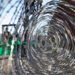 Primer plano a través de un alambre de púas gris enmarañado, colocado en el muro fronterizo oeste del Puerto de San Luis, en Arizona (Estados Unidos). En el fondo, un montacargas verde sostiene una plataforma elevada, sobre la cual dos marines del ejército estadounidense trabajan en la instalación del alambre. También se distingue la valla fronteriza de color marrón bajo un cielo despejado y soleado. Trump