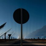 Vista de la central termosolar Gemasolar en Fuentes de Andalucía (España), con su extenso campo de espejos reflectantes y heliostatos inclinados hacia el cielo. En primer plano, una señal de tráfico circular obstruye parcialmente la vista. Transición justa