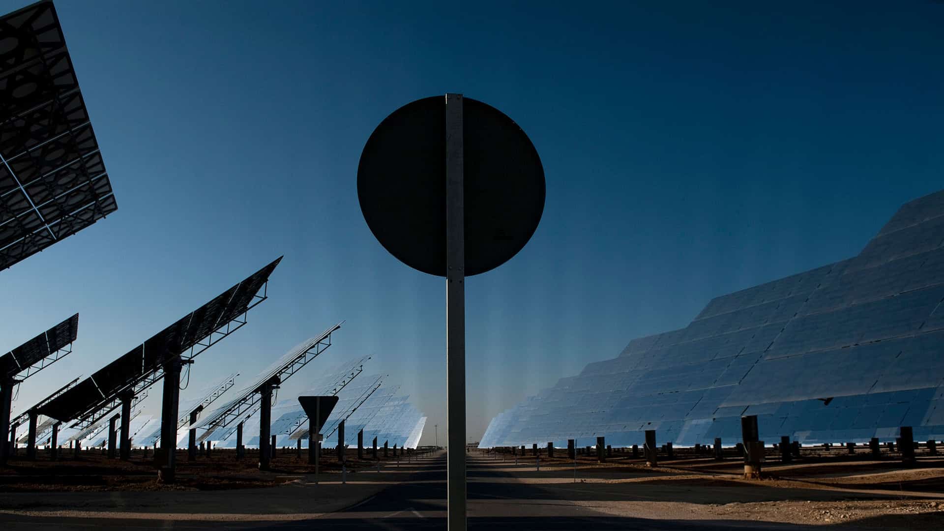 Vista de la central termosolar Gemasolar en Fuentes de Andalucía (España), con su extenso campo de espejos reflectantes y heliostatos inclinados hacia el cielo. En primer plano, una señal de tráfico circular obstruye parcialmente la vista. Transición justa