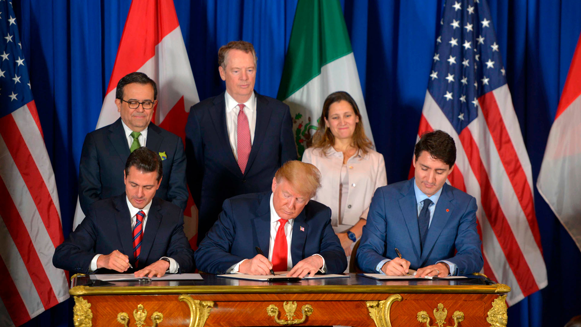 De izquierda a derecha, sentados y firmando el tratado de libre comercio entre EEUU, Canadá y México (USMCA) en la cumbre del G20 en Argentina en el año 2018: Enrique Peña Nieto, presidente de México, con un traje negro, camisa blanca y corbata a rayas rojas y azules; Donald Trump, presidente de EEUU, con un traje azul oscuro, camisa blanca y corbata roja; y Justin Trudeau, primer ministro de Canadá, con un traje, camisa y corbata en distintas tonalidades de azul. Al fondo, las banderas de Estados Unidos, Canadá y México, así como tres personas que acompañan la firma (dos hombres y una mujer de pie), con un fondo azul.