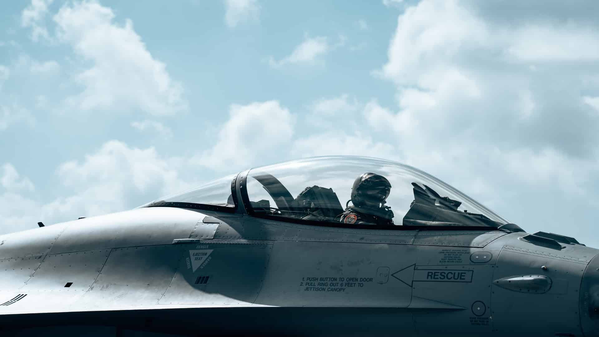 Un piloto belga de F-16, con casco de vuelo, máscara de oxígeno y traje de vuelo completo, está sentado dentro de la cúpula transparente de un F-16 Fighting Falcon de color gris. La aeronave está estacionada en la pista de la Base Aérea de Florennes, bajo un cielo brillante con algunas nubes, durante un ejercicio de Alerta de Reacción Rápida (QRA) de la OTAN. Europa