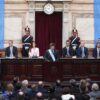 Javier Milei, de pie, con traje oscuro, camisa blanca, corbata negra y la banda presidencial con los colores de la bandera argentina, pronuncia un discurso desde el atril principal del recinto en la 142ª Apertura de Sesiones Ordinarias, en el Congreso de la Nación, en Buenos Aires (Argentina), el 1 de marzo de 2024. Está flanqueado por miembros del gabinete y legisladores. En el fondo, el recinto está decorado con banderas argentinas y se pueden observar los numerosos asistentes en el hemiciclo. Milei