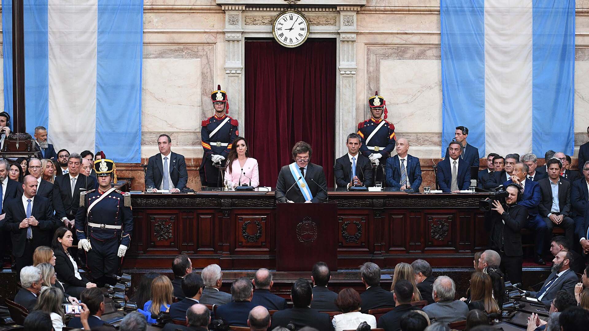Javier Milei, de pie, con traje oscuro, camisa blanca, corbata negra y la banda presidencial con los colores de la bandera argentina, pronuncia un discurso desde el atril principal del recinto en la 142ª Apertura de Sesiones Ordinarias, en el Congreso de la Nación, en Buenos Aires (Argentina), el 1 de marzo de 2024. Está flanqueado por miembros del gabinete y legisladores. En el fondo, el recinto está decorado con banderas argentinas y se pueden observar los numerosos asistentes en el hemiciclo. Milei