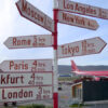 Carteles de flechas en rojo y blanco que muestran direcciones hacia distintas capitales del mundo con el tiempo que toma hacia cada una en las afueras del Aeropuerto de Kangerlussuaq o Søndre Strømfjord en Groenlandia. Al lado de la terminal aérea un avión civil de color rojo de Air Greenland, la principal aerolínea del país.