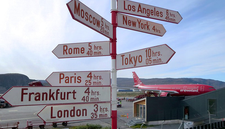 Carteles de flechas en rojo y blanco que muestran direcciones hacia distintas capitales del mundo con el tiempo que toma hacia cada una en las afueras del Aeropuerto de Kangerlussuaq o Søndre Strømfjord en Groenlandia. Al lado de la terminal aérea un avión civil de color rojo de Air Greenland, la principal aerolínea del país.