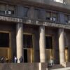 Fachada de la sede del Ministerio de Economía en Buenos Aires, Argentina. Tres personas hacen fila, mientras un policía les observa. Un hombre (con traje y maletín) y una mujer (que camina en sentido contrario) pasan por la acera.