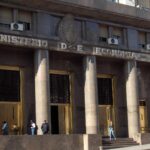 Facade of the Ministry of Economy headquarters in Buenos Aires, Argentina. Three people queue while a policeman watches them. A man (in a suit with a briefcase) and a woman (walking in the opposite direction) pass by on the pavement.