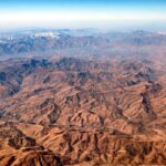 Vista aérea de las montañas de Qandil, en el norte de Irak, que muestra crestas áridas y escarpadas, valles profundos y picos nevados a lo lejos bajo un cielo azul despejado. Un río serpentea por la esquina inferior derecha del paisaje montañoso. Insurgencia kurda