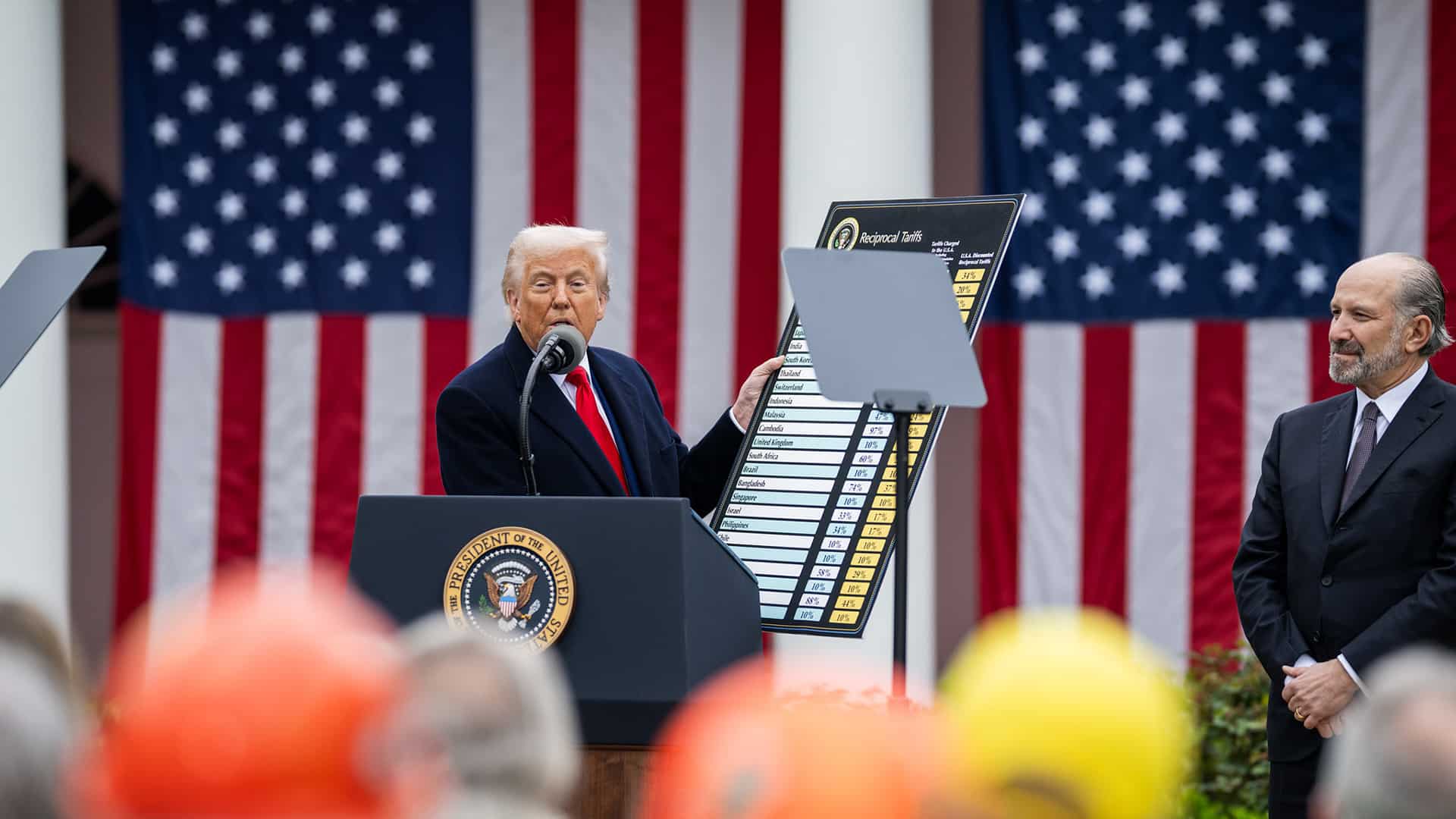 Donald Trump anuncia los aranceles “recíprocos” junto a Howard Lutnick, secretario de Comercio de EEUU (a la derecha), desde un atril con micrófono ante una audiencia congregada en el evento Make America Wealthy Again o “Día de la Liberación” en la Rosaleda de la Casa Blanca, el pasado 2 de abril de 2025. En su mano izquierda, Trump sostiene en alto una tabla con el listado de países y los aranceles (shock arancelario).