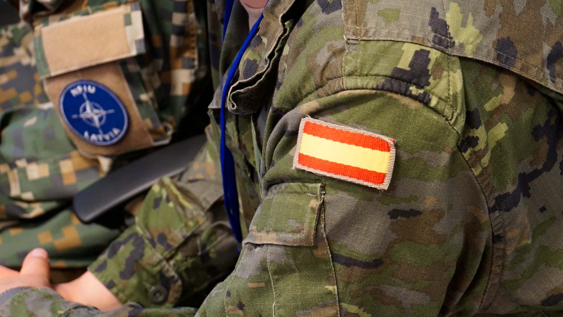 Close-up of two soldiers in camouflage uniforms. One has a Spanish flag patch on their shoulder, and the other has a NATO Latvia patch, representing collaboration within the NATO alliance. Defence