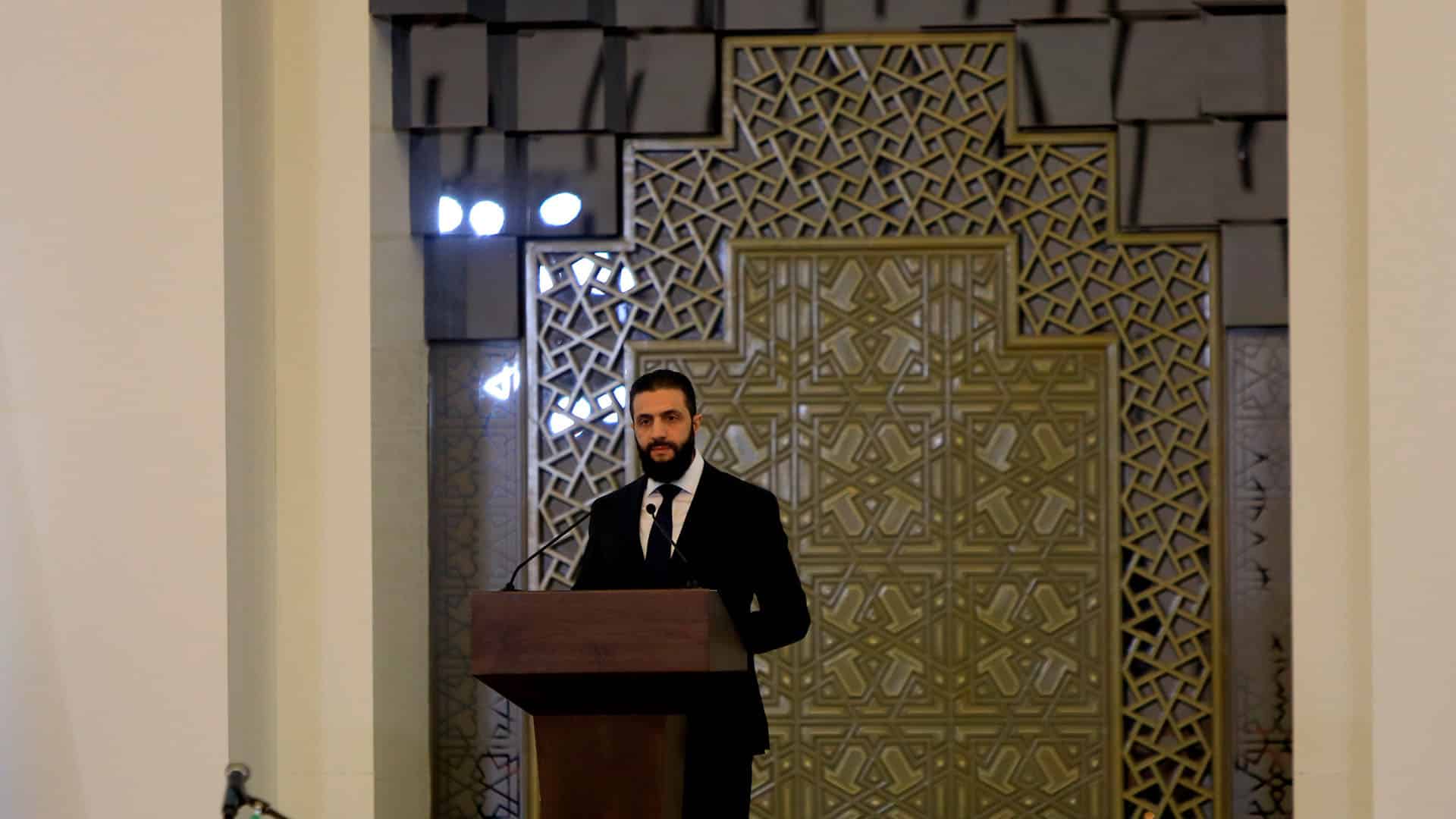 El presidente sirio Ahmad al-Sharaa, de pie, con traje oscuro, camisa blanca y corbata oscura, habla desde un podio de madera durante la investidura de su primer gabinete, frente a banderas y un público reunido en el Palacio del Pueblo (Damasco), el 29 de marzo de 2025. Siria