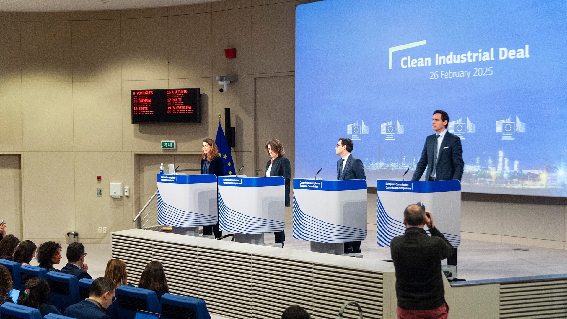 Conferencia de prensa en la sede de la Comisión Europea en Bruselas, el 26 de febrero de 2025. Teresa Ribera, Stéphane Séjourné y Wopke Hoekstra, están de pie detrás de atriles con el logotipo de la Comisión. Una gran pantalla azul al fondo muestra el título “Clean Industrial Deal – 26 February 2025”. En la sala, varios periodistas están sentados tomando notas y fotografías. Clean Industral Deal