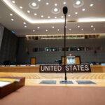 Micrófonos y placas identificativas en inglés de Estados Unidos y Argelia en el Salón vacío del Consejo de Seguridad de las Naciones Unidas, con una iluminación led en el techo, la mesa central en forma de herradura, asientos rojos y azules, y un gran mural en la pared.