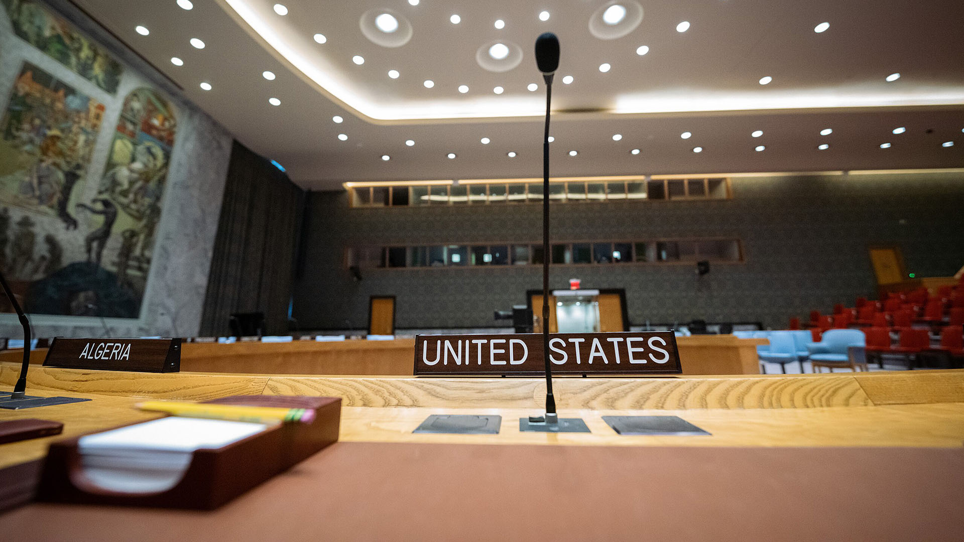 Microphone and nameplate for the United States at a United Nations Security Council meeting room, with the Algeria nameplate visible beside it. The room features modern lighting, a central horseshoe-shaped table, red and blue seating, and a large mural on the wall. Trump