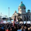 Manifestación organizada por el gobierno de Serbia, el 12 de abril de 2025, bajo el lema “Asamblea popular: no entregamos Serbia”. Al fondo se ve el edificio del Parlamento de Serbia con su cúpula verde, mientras numerosas banderas serbias ondean entre la multitud bajo un cielo despejado. Serbia
