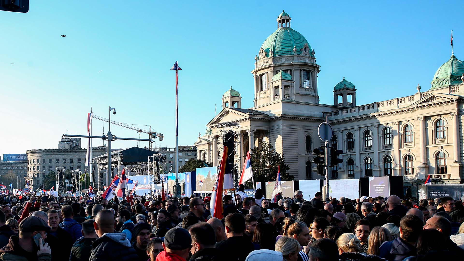 Manifestación organizada por el gobierno de Serbia, el 12 de abril de 2025, bajo el lema “Asamblea popular: no entregamos Serbia”. Al fondo se ve el edificio del Parlamento de Serbia con su cúpula verde, mientras numerosas banderas serbias ondean entre la multitud bajo un cielo despejado. Serbia