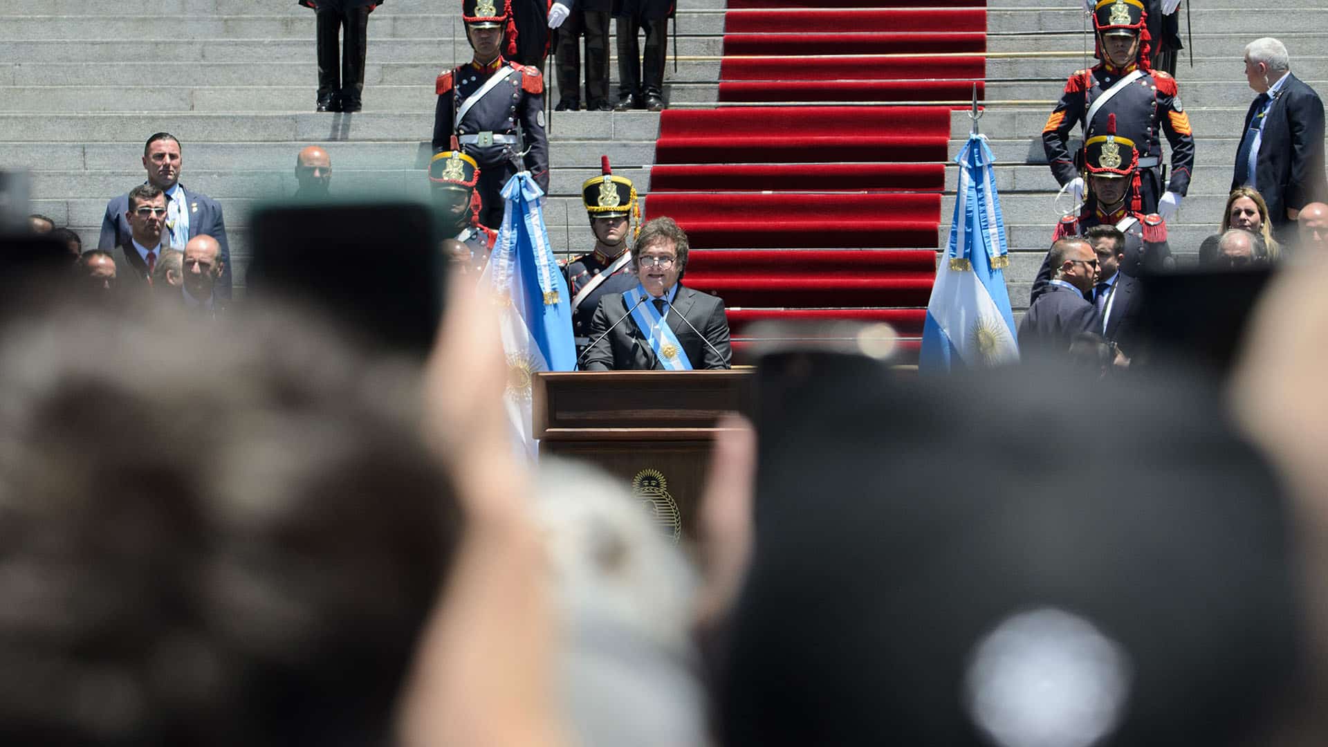 Javier Milei tras un podio mientras pronuncia su discurso frente al Congreso Nacional durante su investidura en 2023. Lleva la banda presidencial y la medalla, flanqueado por banderas argentinas y escoltado por miembros de la Guardia de Granaderos. A sus espaldas se extiende una alfombra roja sobre las escalinatas del Congreso, con numerosos asistentes y personal de seguridad alrededor.