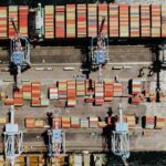 Aerial view of an industrial seaport in Naples, with several cargo cranes lined up and multicoloured shipping containers organised on the quay and aboard a docked cargo ship. Global