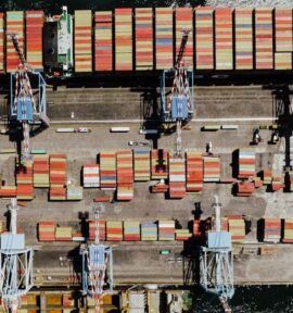 Aerial view of an industrial seaport in Naples, with several cargo cranes lined up and multicoloured shipping containers organised on the quay and aboard a docked cargo ship. Global