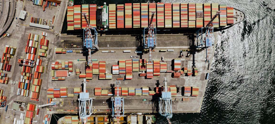 Vista aérea de un puerto marítimo industrial en Nápoles, con varias grúas de carga alineadas y contenedores de transporte multicolores organizados sobre el muelle y a bordo de un buque carguero atracado. UE