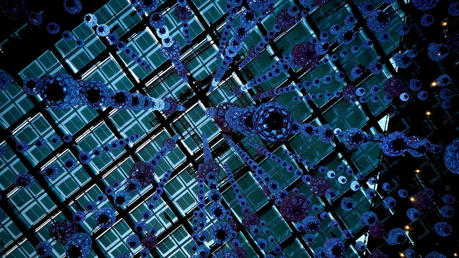 Vista desde abajo de una instalación artística luminosa con formas esféricas y cilíndricas en diferentes tonos de azul y verde, suspendidas en el aire contra un techo enrejado de vidrio. Tecnologías cuánticas