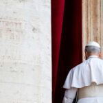 El papa León XIV, visto de espaldas y vestido con la sotana papal, se aleja tras pronunciar su primera oración del Regina Caeli desde un balcón con vistas a la plaza de San Pedro. A la izquierda se aprecia parte del edificio revestido de travertino claro, mientras que detrás de él se alza una columna del mismo material, enmarcada por cortinas rojas. Papa